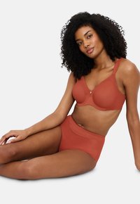 Ženska s kodrasto lasmi sedi, oblečena v usklajen terrakotasti modrček in hlačke visoko pasu, na enobarvni svetli ozadju.