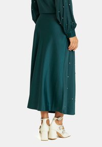 Largo vestido de satén color teal con acentos de perlas a lo largo del costado, combinado con zapatos de tacón blancos que presentan hebillas decorativas y un diseño de corte.