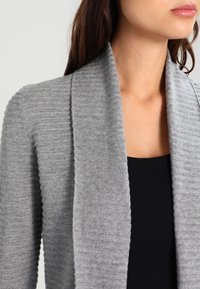 Cardigan grigio con un motivo a coste testurizzato, design aperto davanti e materiale in maglia morbido. Indossato sopra una maglietta nera, che mette in mostra il colletto.