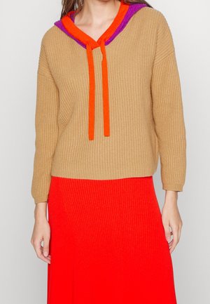 Femme portant un pull côtelé beige avec une capuche violette et orange et des liens, associé à une jupe côtelée rouge vif.