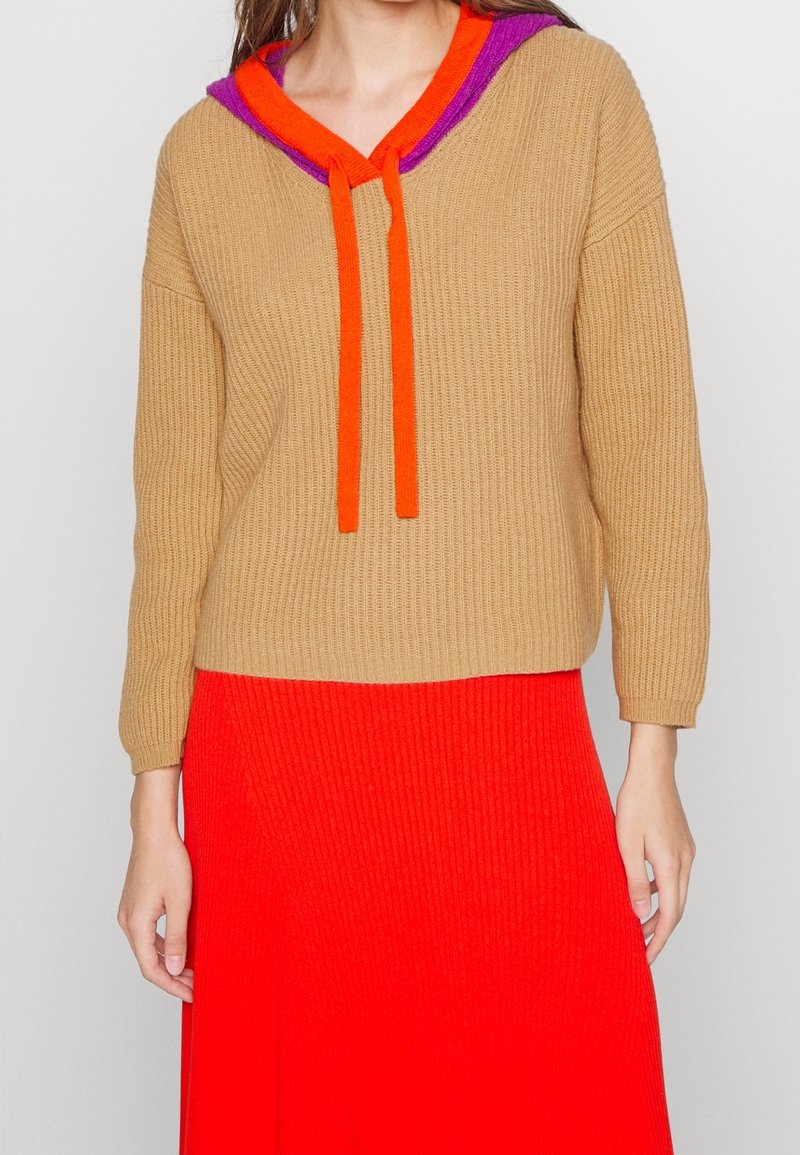 Femme portant un pull côtelé beige avec une capuche violette et orange et des liens, associé à une jupe côtelée rouge vif.