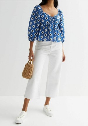 Femme portant un chemisier bleu à motifs, un pantalon blanc ample et court, des baskets blanches, et tenant un petit sac tissé.