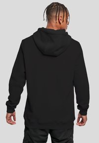 Homme avec des cheveux tressés portant un sweat à capuche noir uni et un pantalon sombre, de dos contre un fond gris.