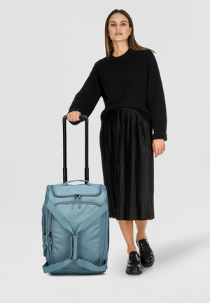 Ein hellblauer Rollkoffer mit doppelten Reißverschlüssen und Seitentaschen, ausgestattet mit einem stabilen Griff und Kreuzgurten zur Sicherung. Texturiertes Material.