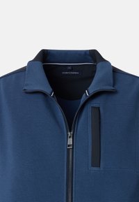 Giacca a zip navy con superficie testurizzata, colletto rialzato e dettaglio della tasca rettangolare. Presenta accenti neri a contrasto e un'etichetta del brand.