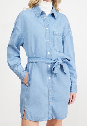 Femme portant une robe chemise en denim bleu clair avec ceinture à la taille, poche poitrine et manches longues, par-dessus un col roulé blanc.