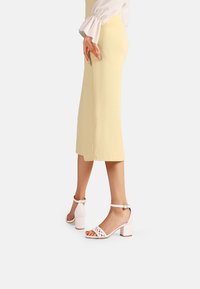 Sandali bianchi con tacco a blocco presentano un motivo a taglio sulla cinghia e una fibbia alla caviglia. Indossati con un vestito giallo, evidenziano la parte inferiore della gamba.