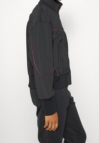 Giacca sportiva nera con colletto alto, accenti di cuciture rosse, tasca con zip e polsini a coste. Texture morbida, design aderente.