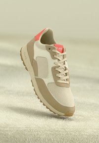 Beige sneaker with mesh and suede materials, featuring a white panel and a red accent at the heel. Includes flat laces and a rubber sole.