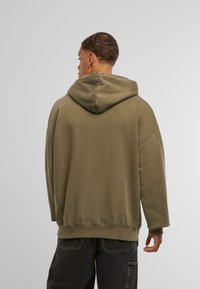 Oliņzaļš kapuce, izgatavota no mīksta auduma, ar plašu kapuci, nolaižamām plecu daļām un ribotām piedurknēm un apakšmalu. Minimālistisks dizains, bez rakstiem.