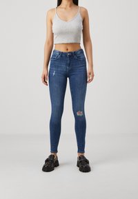 Jeans skinny de mezclilla azul oscuro con talle alto, que presentan detalles desgastados y material elástico. Combinados con una camiseta sin mangas gris corta y zapatos negros.