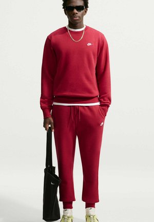 Mann trägt roten Nike Trainingsanzug, weiße Socken, gelbe Sneaker, schwarze Sonnenbrille, goldene Kette, hält eine schwarze Nike Tragetasche vor einem einfarbigen Hintergrund.