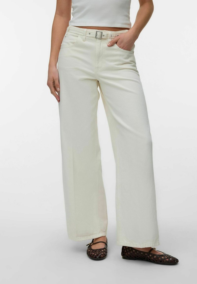Femme portant un pantalon large crème avec une ceinture, un crop top blanc et des ballerines tissées noires, debout avec une main dans la poche.