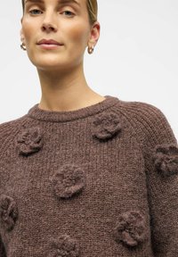 Rudas megztinis su tekstūru, puošnus pakeltomis gėlėmis. Apvalus iškirpimas ir ilgos rankovės. Minkšta, storo audinio medžiaga.