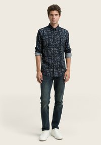 Marineblauwe shirt met geometrische patronen, lange mouwen opgerold bij de manchetten, gecombineerd met donkere jeans en witte sneakers.