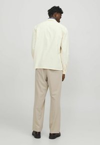 Chemise beige clair à manches longues, en coton, avec un ourlet arrondi, assortie à un pantalon beige clair et des chaussures foncées. Design minimaliste.