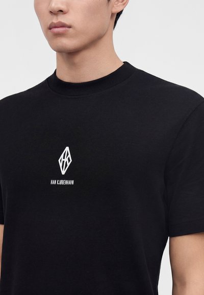 Άντρας που φοράει μαύρο T-shirt με λευκό γεωμετρικό λογότυπο και κείμενο "HAN KJØBENHAVN" στο στήθος σε απλό φόντο.