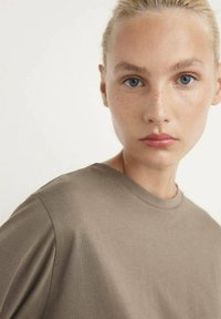 Bruine katoenen t-shirt met een ronde halslijn en korte mouwen, met een gladde textuur en een relaxte pasvorm. Huidtint is licht met sproeten.