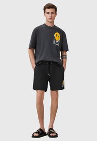 T-shirt nera con grafica, design a faccina sorridente gialla e testo bianco, abbinata a pantaloni corti neri e sandali neri.