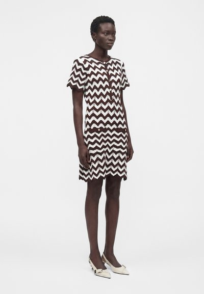 Femme debout portant une robe à manches courtes, longueur genoux, avec un motif zigzag marron et blanc et des chaussures beige à bouts pointus sur un fond uni.