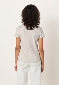 Žena s kučeravými vlasmi stojí chrbtom k nám, oblečená v svetlosivej krátkokrkej košeli a svetlomodrých džínsoch na jednoduchom pozadí.