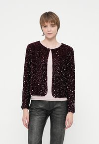 Jaunas žmogus su trumpais plaukais vilki tamsiai purpurinį blizgučių švarką virš šviesiai rožinės spalvos marškinėlių ir juodų tekstūruotų kelnių prieš paprastą foną.