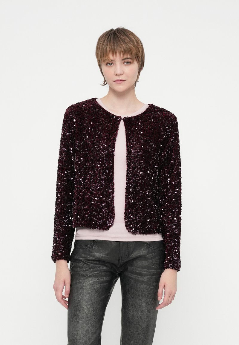 Jaunas žmogus su trumpais plaukais vilki tamsiai purpurinį blizgučių švarką virš šviesiai rožinės spalvos marškinėlių ir juodų tekstūruotų kelnių prieš paprastą foną.