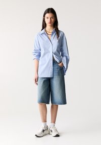 Ljust blå randig skjorta med långa ärmar, avslappnad passform, tillsammans med knälånga denimshorts och vita sneakers med svarta detaljer.
