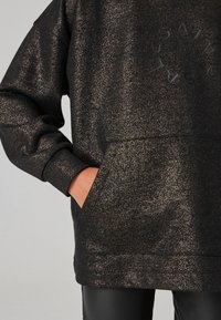 Sudadera negra con destellos metálicos, presenta un bolsillo frontal y un escote redondeado. Muestra un logo circular en el centro.