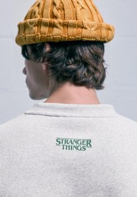 Persona con capelli castani che indossa un berretto di lana giallo e una maglietta grigia chiara con testo verde "Stranger Things" sul colletto posteriore.