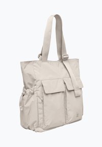 Sac fourre-tout beige clair en tissu durable, avec deux poches à rabat à l'avant, des bretelles réglables et des poches en mesh sur les côtés pour un rangement supplémentaire.