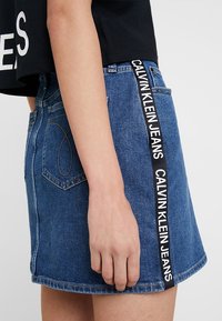 Person som bär en blå denimminikjol med "Calvin Klein Jeans" tryckt på en svart sidremsa, kombinerat med en kort svart topp.