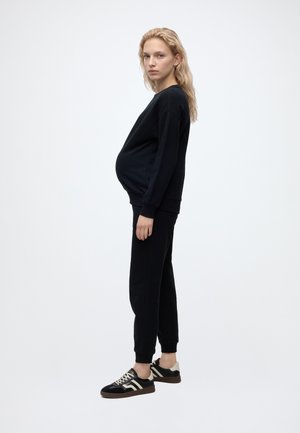 Femme enceinte aux longs cheveux blonds, vêtue d’un sweat-shirt noir et d’un pantalon, debout de profil, portant des baskets noires et blanches sur un fond uni.