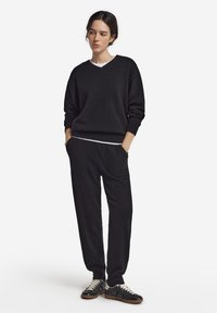 Svart v-ringad tröja med ribbade muddar, kombinerad med svarta joggerbyxor och svarta sneakers med vita snören och gummisulor.