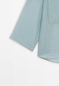 Chemise à manches longues en tissu bleu clair froissé avec une poche poitrine, montrée partiellement de la manche et du corps sur un fond blanc.