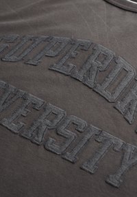 Camiseta gris oscuro con letras bordadas en relieve "UNIVERSIDAD SUPERIOR" en un tono contrastante, con un acabado de tejido suave y texturizado.