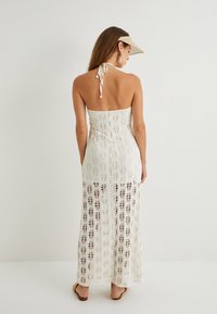 Langes weißes Häkelkleid mit offenen Mustern, rückenfreiem Design und Halterneck-Bindung. Kombiniert mit einer Visor und minimalistischen Accessoires.