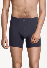 Manlig bål och ben iförda mörkgrå boxertrosor, stående framför en vit bakgrund med avslappnade armar.