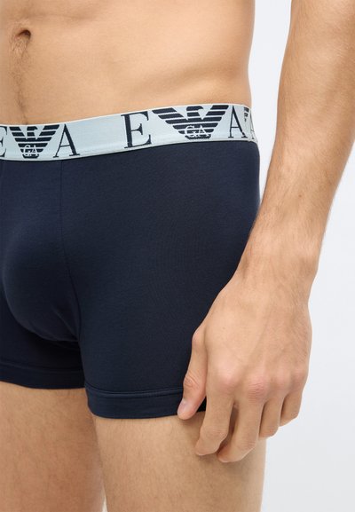 Tengerészkék pamut férfi boxeralsó világoskék elasztikus derékpánttal, amelyen fekete logónyomat található. Simaságú anyag és testhezálló kialakítás.