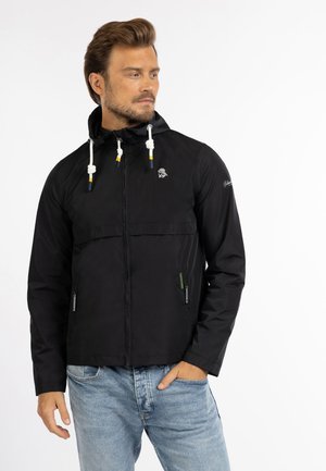 Uomo che indossa una giacca nera con cappuccio e cordini bianchi e jeans azzurri chiari, in piedi davanti a uno sfondo bianco uniforme.