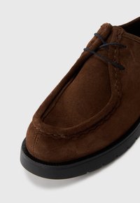 Chaussure en daim marron avec un bout arrondi, semelle en caoutchouc noir et couture contrastante. Dispose d'un système de laçage et d'une texture douce.