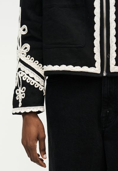 Κοντινό πλάνο σε μανίκι και κορμό μαύρου σακακιού με λευκή κεντητή δαντέλα και ασημένιο φερμουάρ, φορεμένο με μαύρο παντελόνι.