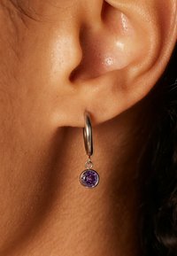 Zilveren oorring met een ronde, geplaate paarse edelsteen hanger. De edelsteen is geplaatst in een gepolijste zilveren zetting. Close-up van de huid.