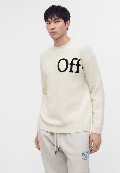 Jeune homme portant un pull crème avec un texte partiel "Off" et un pantalon de survêtement gris clair avec un logo bleu et des détails à cordon.