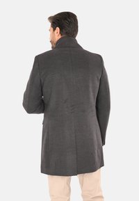 Uomo che indossa un cappotto su misura grigio scuro visto di spalle, abbinato a pantaloni beige chiaro su uno sfondo bianco uniforme.