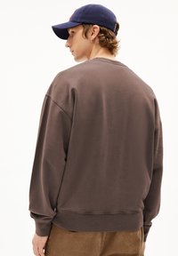 Sweatshirt marron avec une coupe décontractée, poignets et encolure côtelés. Porté avec une casquette sombre ; un pantalon en côtes de velours texturé est visible en dessous.