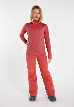 Rotes langärmliges Oberteil mit halbem Reißverschluss-Kragen, kombiniert mit locker sitzenden roten Hosen aus Strukturstoff. Schwarze Schuhe runden das Outfit ab.