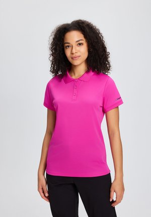 Kobieta z kręconymi włosami, ubrana w jasnoróżową koszulkę polo z krótkim rękawem i czarne spodnie, na jednolitym jasnym tle.