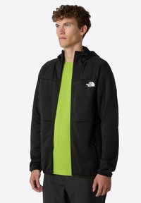 Schwarze Kapuzenjacke mit Reißverschlusstaschen und einer weichen Textur, kombiniert mit einem leuchtend grünen T-Shirt darunter; mit Logo auf der Brust.