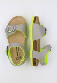 Sandales grises avec des accents jaunes vifs, boucle réglable, semelle en liège et semelle en caoutchouc texturé. Elles disposent de deux sangles pour un maintien sécurisé.